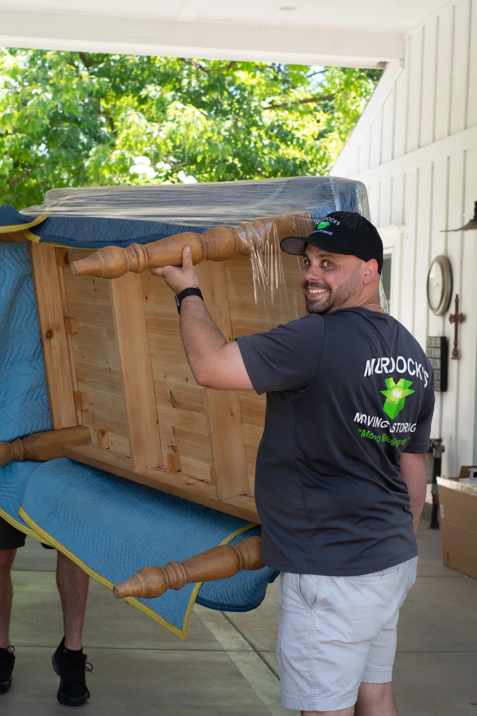Smiling Murdock's Moving & Storage mover in branded shirt and cap carries a plastic-wrapped wooden table