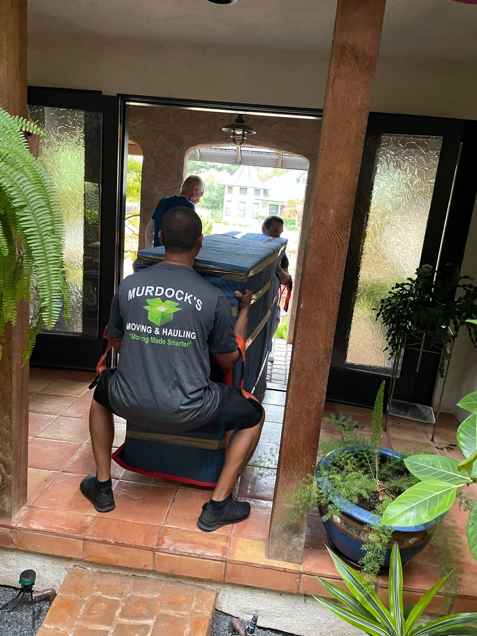 Murdock's Moving & Storage crew carry a large blanket-wrapped item through a tiled entryway using harness straps