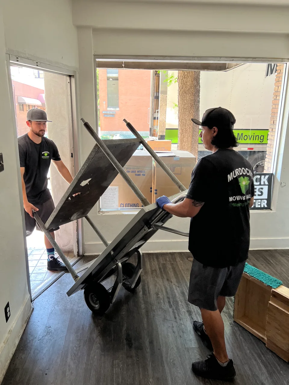 Two Murdock's Moving & Storage movers in branded shirts carry a table on a hand truck through a doorway