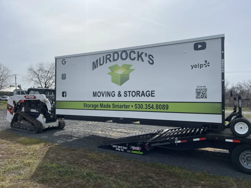 Murdock's Moving & Storage MODS portable storage container beside a flatbed trailer with loading ramp in a driveway