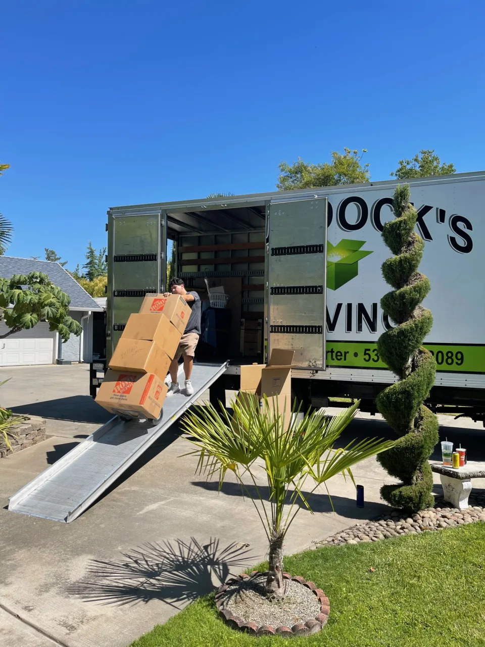 Murdock's Moving & Storage mover wheels a stack of packed boxes down the loading ramp of a branded truck