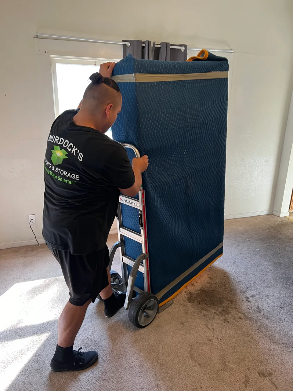 Murdock's Moving & Storage mover wheels a blue blanket-wrapped dresser on a hand truck indoors