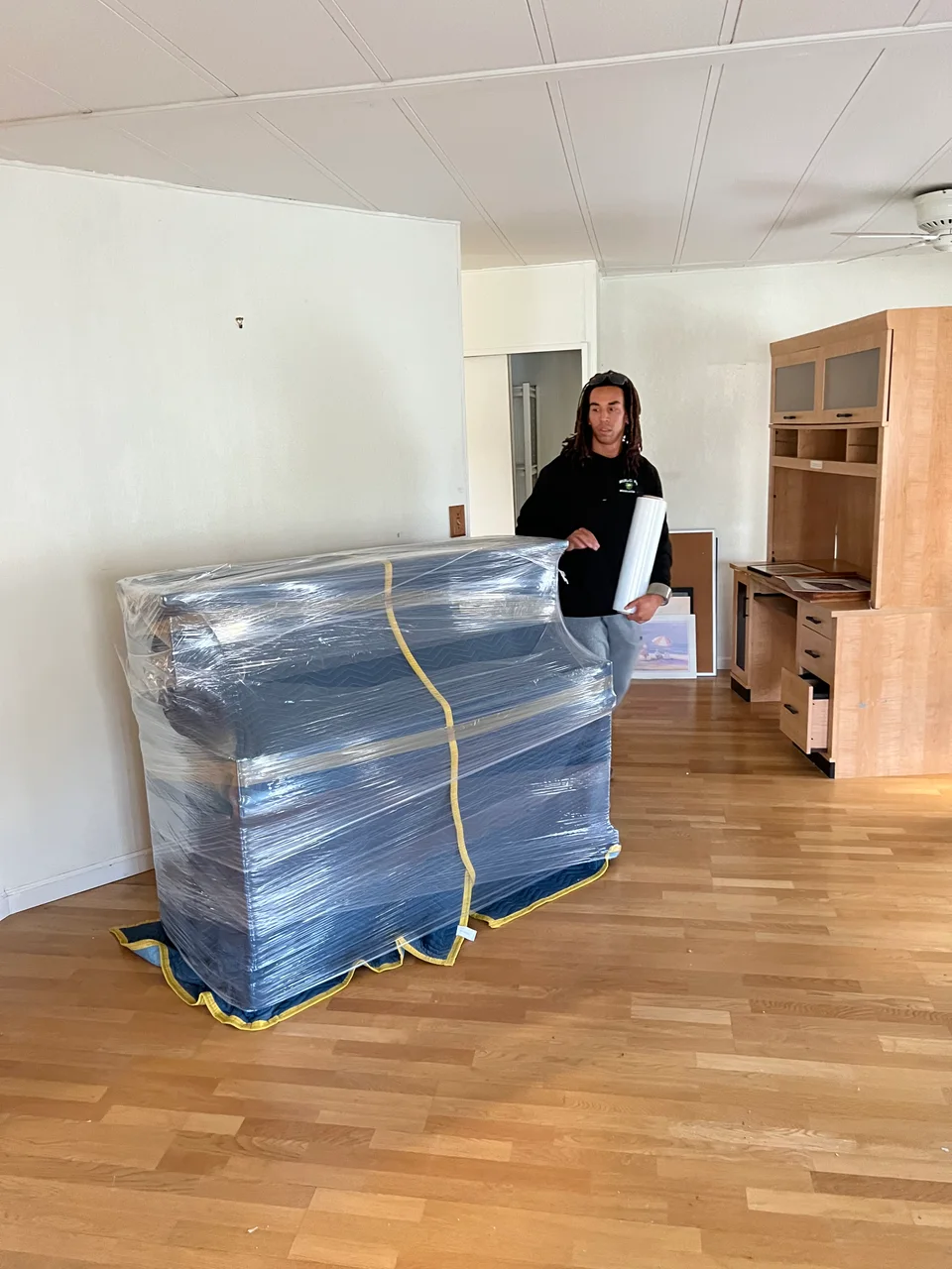 Murdock's mover with dreadlocks applying stretch shrink wrap over a blue-padded, blanket-covered large item indoors