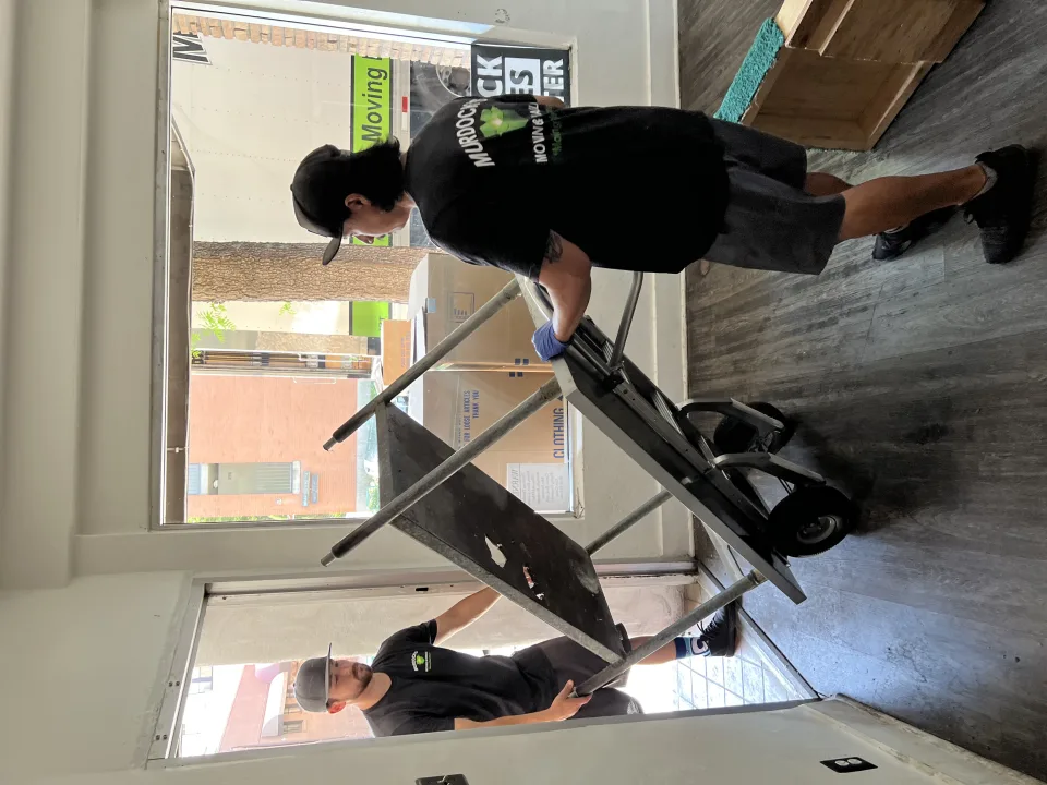 Two Murdock's Moving crew members carry padded, shrink-wrapped table legs and top alongside the branded box truck