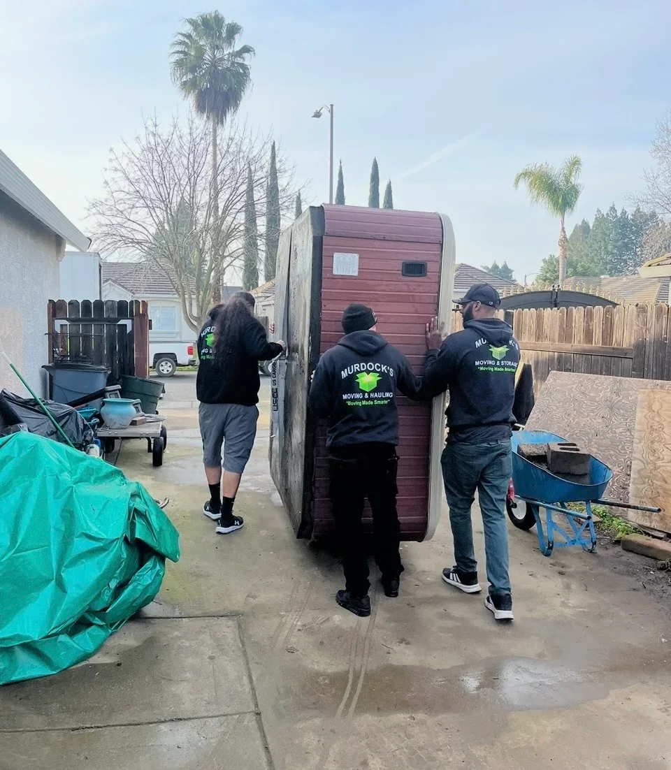 Three Murdock's Moving & Hauling crew members maneuver a hot tub on its side through a backyard