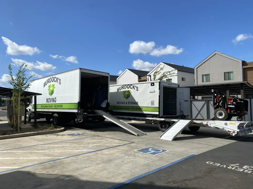 Murdock's Moving box truck beside a branded MOD storage container on a trailer with a loading tractor under blue sky