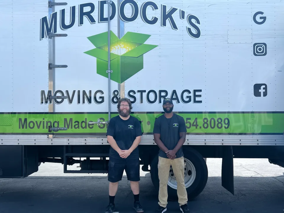 Two smiling Murdock's Moving & Storage crew members standing in front of the large branded truck